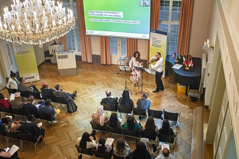 Cornelia Pfeil und Cornelius Köhler spielen Geige vor einer Kulisse mit Flaggen verschiedener Länder, einige Zuhörende sind im Vordergrund sichtbar.