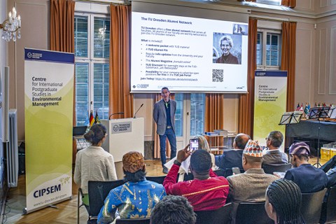 Jeremiah Peterson spricht zu den Teilnehmenden der Veranstaltung, im Vordergund ist Publikum zu sehen, im Hintergrund eine Präsentation..