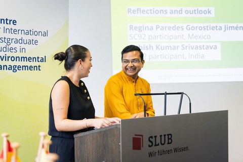 Foto zeigt Regina Paredes Gorostieta Jimnez und Ashish Kumar Srivastava am Rednerpult. Beide lachen einander an, während sie die Kursteilnehmenden während der Abschlussfeier des SC92 ansprechen.