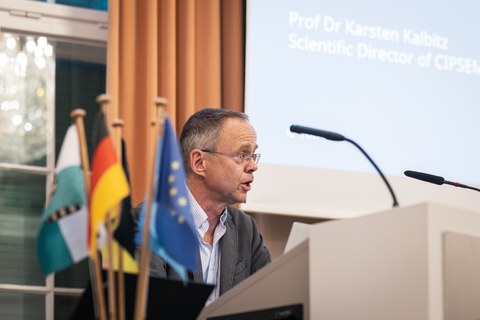 Prof. Karsten Kalbitz, Wissenschaftlicher Direktor von CIPSEM, spricht hinter einem Rednerpult im Festsaal des Rektorats der TU Dresden. Neben ihm sind mehrere Flaggen zu sehen.