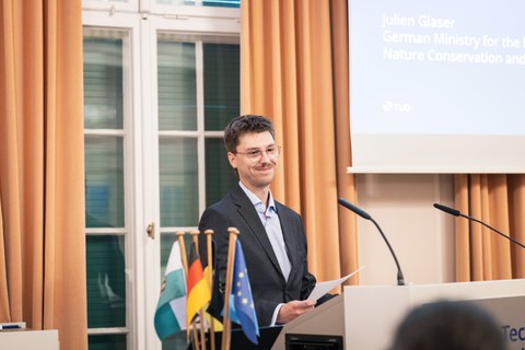 Julien Glaser vom Bundesministerium für Umwelt, Klimaschutz, Naturschutz und nukleare Sicherheit lächelt hinter einem Rednerpult im Festsaal des Rektorats der TU Dresden.