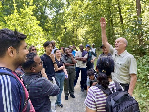 Gruppe internationaler Weiterbildungsteilnehmender auf einem Waldweg, ein Mann in forstlicher Kleidung erklärt.
