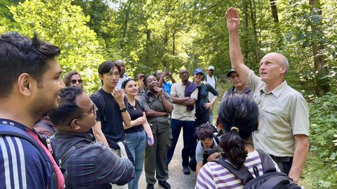 Gruppe internationaler Weiterbildungsteilnehmender auf einem Waldweg, ein Mann in forstlicher Kleidung erklärt.