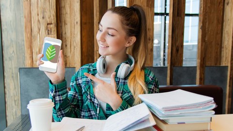 Frau, Studentin, Schreibtisch, Bücher, Handy im Hand, Frau zeigt auf ihren Handybildschirm