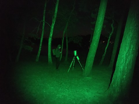 The photo shows a laser scanner with a person standing in front of it, with trees all around. It is a night shot