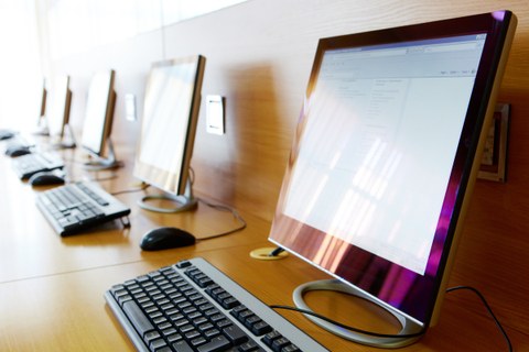 mehrere Computerbildschirme mit Tastaturen, aufgereiht auf einem langen Tisch vor einer Wand