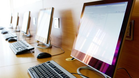 mehrere Computerbildschirme mit Tastaturen, aufgereiht auf einem langen Tisch vor einer Wand