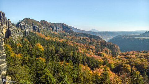 Vom linken Bildrand ziehen sich Sandsteinformationen (Schrammsteingrat) in die Bildmitte in Richtung Hintergrund. Der Vordergrund zeigt herbstlich gefärbten Wald und im rechten Drittel das etwas versteckte Elbtal mit einem Stück Elbe und steiler Talflanke