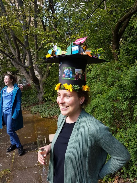 laughing woman with a doctor hat and a glass of champagne in her hand stands in a park. In the background is another person. 