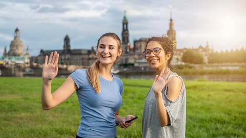 Foto zeigt zwei lächelnde und winkende Studentinnen an der Elbe.
