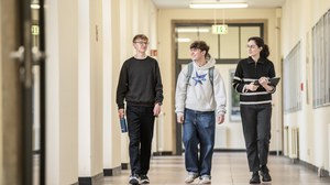 Drei junge Personen laufen sich unterhaltend einen Gang entlang auf die Kamera zu. Die rechte Person hält dabei Unterlagen in den Händen und die linke trägt eine Trinkflasche.
