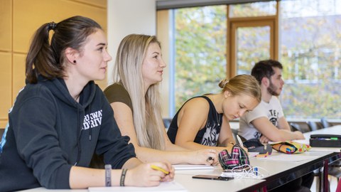 Vier Studenten sitzen in einem Seminarraum. Drei schauen nach vorn und eine schreibt.
