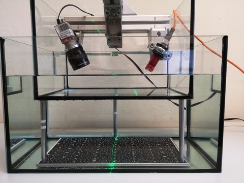 Versuchsaufbau eines Lasertriangulationssystems. Ein Linienlaser befindet sich in einem wasserdichten Glasgehäuse und strahlt eine gebogene Linie auf eine Oberfläche, die sich unter Wasser befindet. Zusätzlich ist eine Kamera im Glasgehäuse.