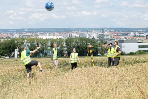 Gruppe von fünf Studierenden der Geodäsie auf Feld, spielen mit Wasserball, Erdmotiv, im Bildzentrum Vermessungsinstrument