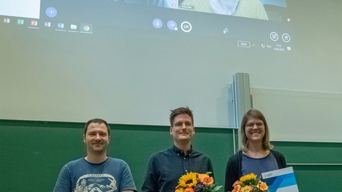 Foto. Drei Personen stehen nebeneinander vor einer Tafel, lächeln und halten Blumensträuße und Urkunden in den Händen. Im Hintergrund ist die Projektion einer Videoübertragung von zwei weiteren lächelnden Personen zu sehen.