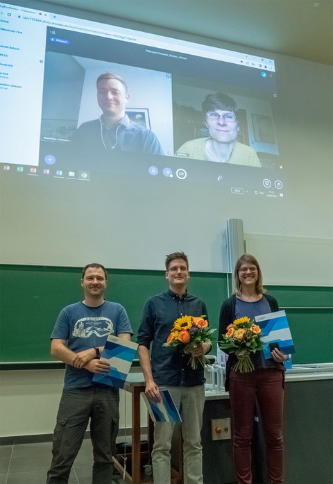 Foto. Drei Personen stehen nebeneinander vor einer Tafel, lächeln und halten Blumensträuße und Urkunden in den Händen. Im Hintergrund ist die Projektion einer Videoübertragung von zwei weiteren lächelnden Personen zu sehen.