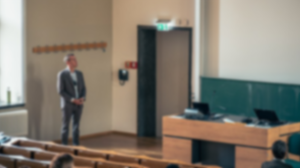 weichgezeichnetes Foto einer Vorlesung, bei der der Saal zu erkennen ist sowie eine Lehrperson