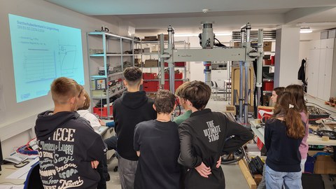 Eine andere Schülergruppe steht im Bahnlabor der Professur für schienenfahrwege. Sie stehen mit dem Rücken zur Kamera und verfolgen eine Präsentation zum Thema Durchschubwiderstand