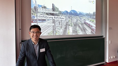 Professor Liu steht am Pult im Vorlesungssaal POT 361. Hinter ihm an der Wand ist das Startbild der Vorlesungsreihe geöffnet.