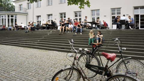 This picture shows two bicycles in the right foreground and the TU Dresden Altmensa in the background with several students sitting on the stairs, having lunch and conversing.