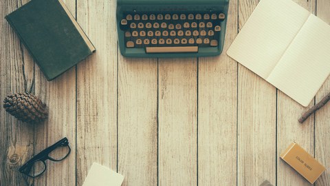This picture shows various items on a table, including a typewriter in the center surrounded by a pad of blank paper, a closed and an open notebook, a pinecone, eye glasses, a small stick, an eraser, and an empty ink well.