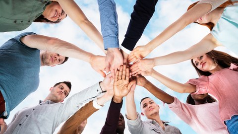 Twelve people standing in a circle place their hands on top of each other in the middle of the circle. 