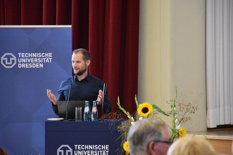 Dr. Sven Lißner hält einen Vortrag, im Hintergrund Logo der TU