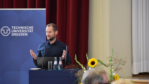 Dr. Sven Lißner hält einen Vortrag, im Hintergrund Logo der TU