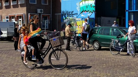 Zu sehen ist eine Straßenszene in der eine Frau auf dem Fahrrad mit Kindersitzen fährt