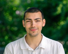 Auf dem Bild sieht man einen Mann. Er trägt ein weißes Polohemd, hat kurze schwarze Haare und lächelt in die Kamera.