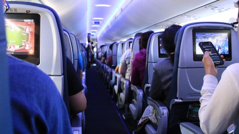 Foto: Innenraum eines Flugzeugs. Blick den Gang entlang. Links und rechts sitzen Passagiere. Einige schauen auf ihr Handy. Andere blicken auf Bildschirme in der Sitzlehne des Platzes vor ihnen.