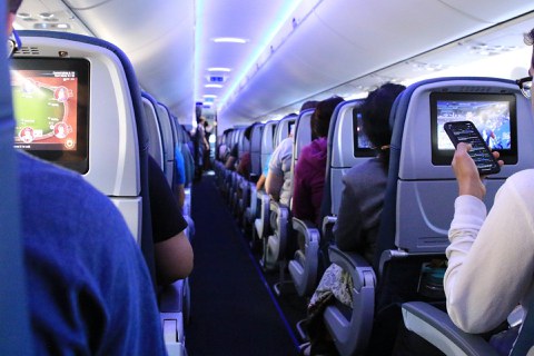 Foto: Innenraum eines Flugzeugs. Blick den Gang entlang. Links und rechts sitzen Passagiere. Einige schauen auf ihr Handy. Andere blicken auf Bildschirme in der Sitzlehne des Platzes vor ihnen.
