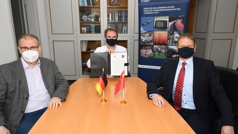 Drei herren sitzen mit Mundschutz an einem Tisch, der mittlere hält einen Vertrag hoch. Auf dem Tisch sind zwei kleine fahnen, die deutsche und die chinesische.