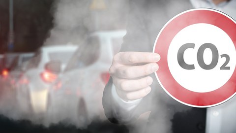 Eine Person hält ein rundes Straßenschild. Es ist Rot umrandet und in der Mitte steht CO2. Im Hintergrun ist ein Stau und Abgase.