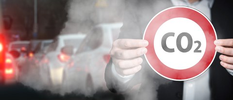 Eine Person hält ein rundes Straßenschild. Es ist Rot umrandet und in der Mitte steht CO2. Im Hintergrun ist ein Stau und Abgase.
