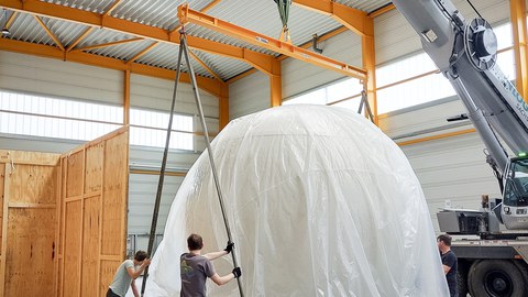 Eine halbrunde Kuppel wird in einer Halle per Kran von einem Lkw gehoben
