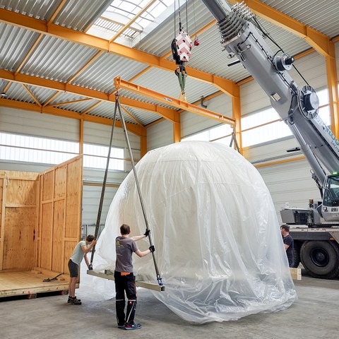 Eine halbrunde Kuppel wird in einer Halle per Kran von einem Lkw gehoben