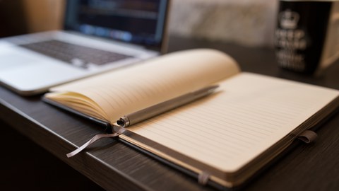 Ein leeres Notizbuch liegt auf einem Tisch, auf dem ein Laptop und eine Tasse steht.