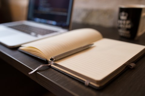 Ein leeres Notizbuch liegt auf einem Tisch, auf dem ein Laptop und eine Tasse steht.