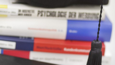 Eine Dissertation liegt neben verschiedenen gestapelten Büchern, auf denen eine Abschlussmütze platziert ist