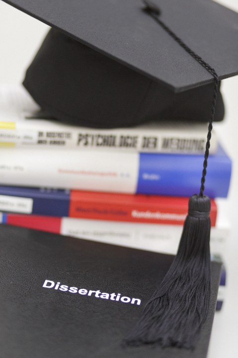 Eine Dissertation liegt neben verschiedenen gestapelten Büchern, auf denen eine Abschlussmütze platziert ist