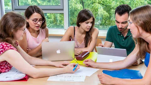 4 Personen sitzen am Tisch und arbeiten gemeinsam mit Zettel + Stift und Laptop