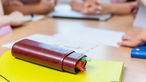 Hefter mit Stiftebox auf dem Tisch, ringsrum sieht man die Hände von Studierenden, die am Tisch sitzen