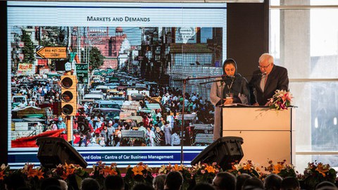 A man and a woman with a headscarf stand on a stage and give a speech to the audience. 