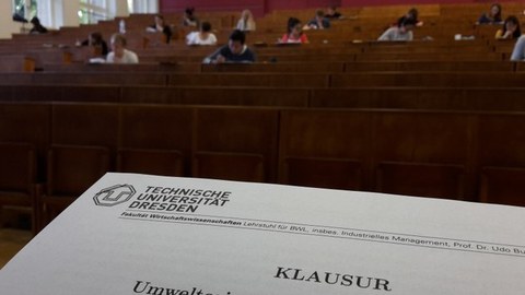 Teil eines Prüfungsbogen im Vordergrund. Im Hintergrund ein Hörsaal mit Studenten die Prüfung schreiben.