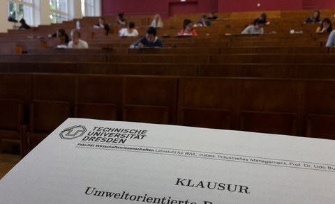 Teil eines Prüfungsbogen im Vordergrund. Im Hintergrund ein Hörsaal mit Studenten die Prüfung schreiben.