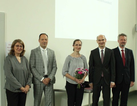 5 Personen befinden sich in einer Reihe. Die Frau in der Mitte hält einen kleinen Blumenstrauß in den Händen.