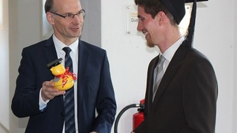 1 man is wearing a doctor hat. 1 man hands over a rubber duck.