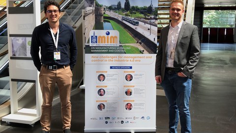 Herr Alarcon und Herr Hoffmann stehen im Konferenzgebäude vor einem Veranstaltungsposter.