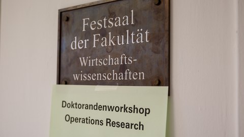 Schild mit der Aufschrift "Festsaal der Fakultät Wirtschaftswissenschaften". Darunter ein Zettel mit der Aufschrift "Doktorandenworkshop Operations Research"."